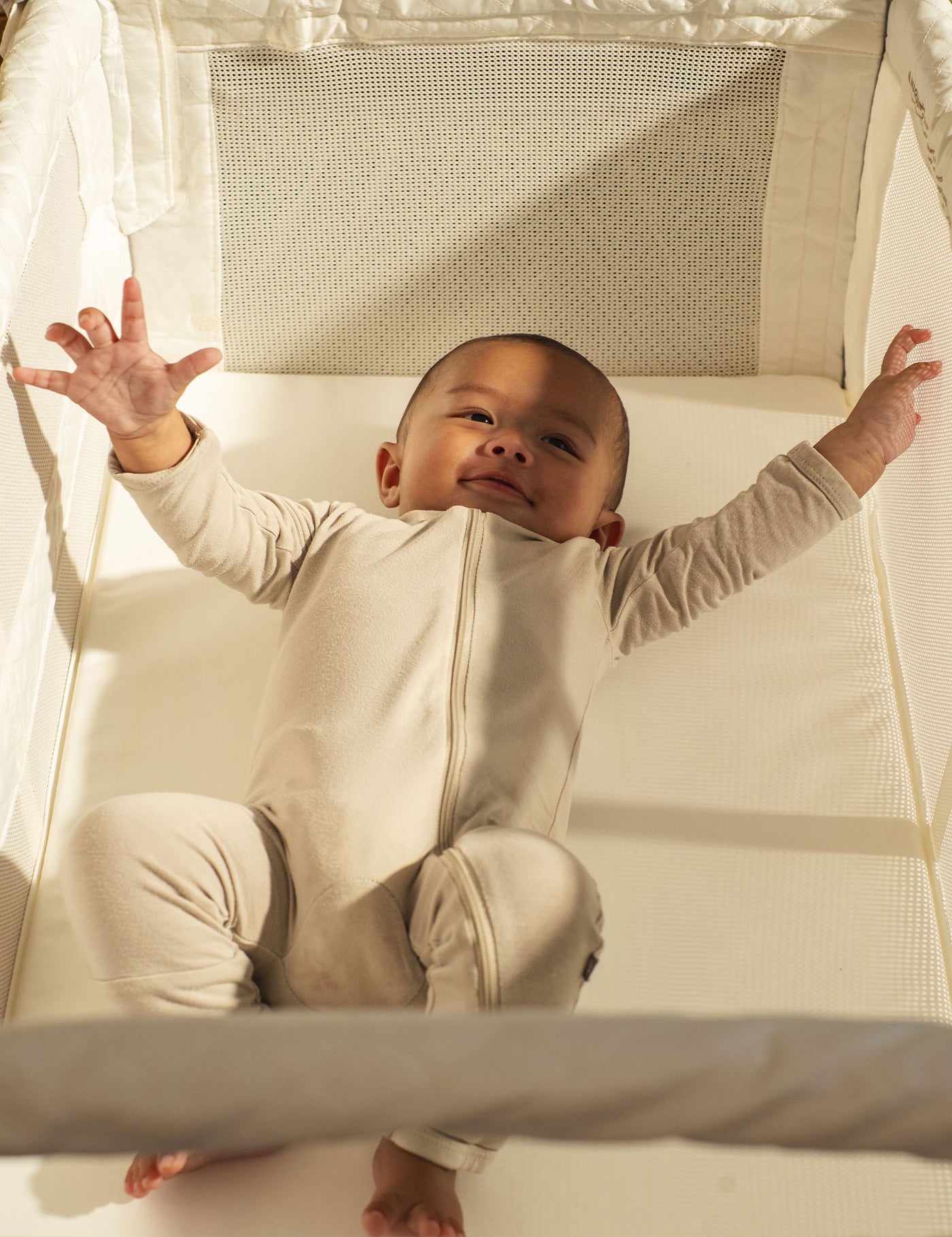 A baby wearing a cream-colored long-sleeve footless onesie sits inside a white Arm's Reach Clear-Vue ™  Co-Sleeper ® bassinet, reaching out with both hands.