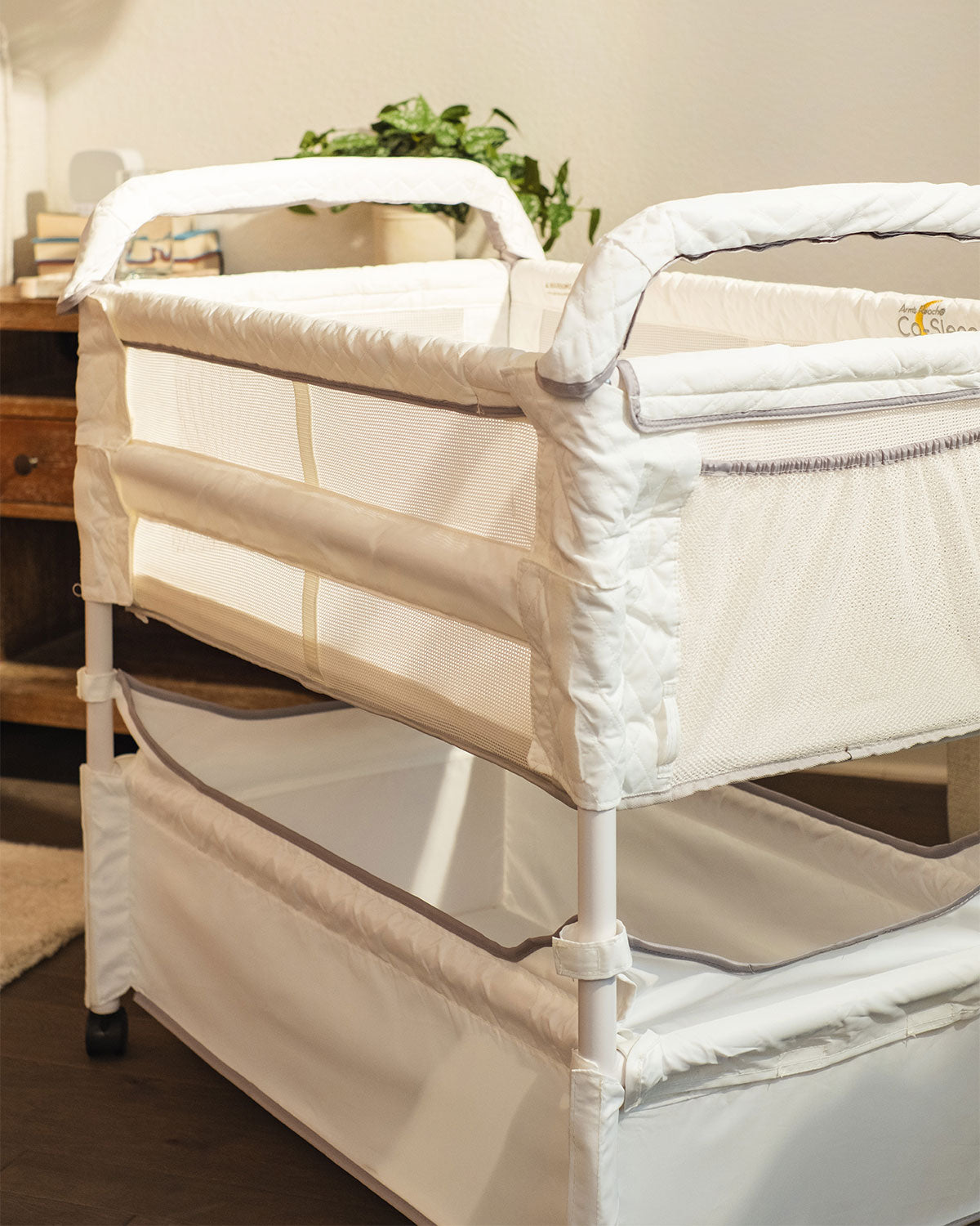 A white Arm's Reach Clear-Vue™ Co-Sleeper® bassinet with padded edges and a mesh side panel, shown in a softly lit nursery setting with a wooden cabinet and plant in the background.