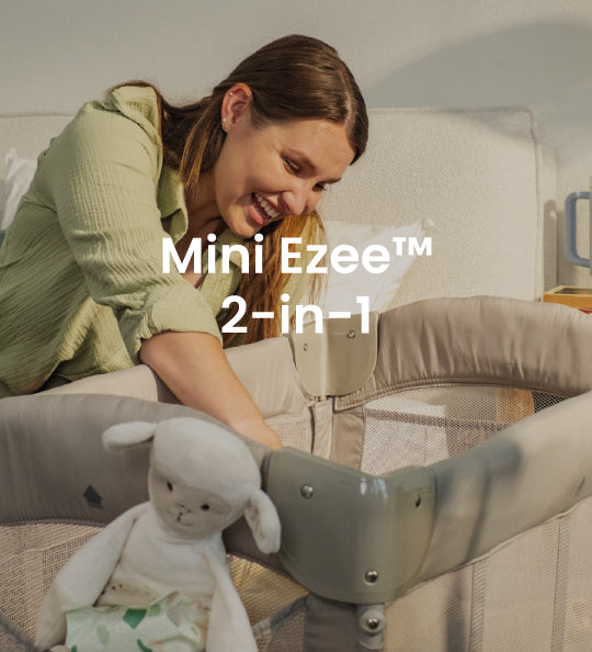 A woman in a light green top leans over a gray and beige baby crib, smiling at a white plush toy inside. White text overlay reads 'Mini Ezee 2-in-1'.