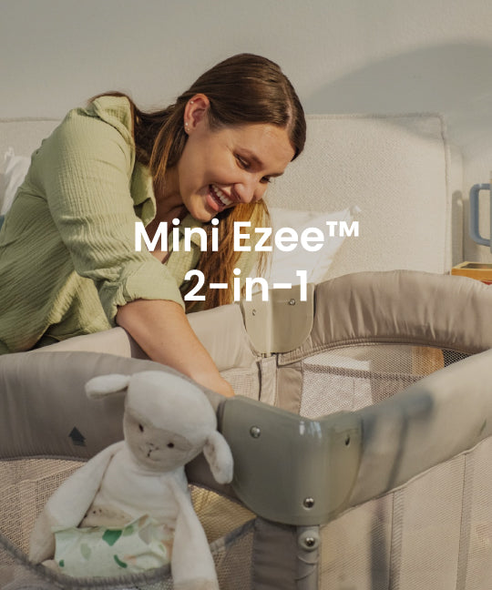 A woman in a light green top leans over a gray and beige baby crib, smiling at a white plush toy inside. White text overlay reads 'Mini Ezee 2-in-1'.