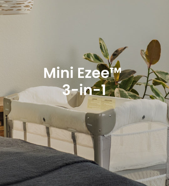 A bassinet with a white mesh side and padded cream interior, shown in a styled room setting with a plant, wall, and blanket. White text overlay reads 'Mini Ezee 3-in-1'.