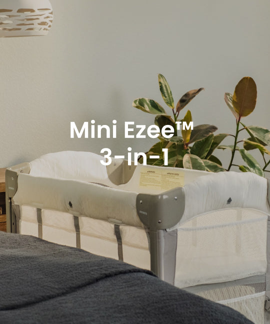 A bassinet with a white mesh side and padded cream interior, shown in a styled room setting with a plant, wall, and blanket. White text overlay reads 'Mini Ezee 3-in-1'.