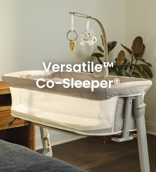 A Versatile Co-Sleeper bassinet in a nursery setting, featuring a white and gray fabric liner, a white metal frame, and an adjustable mobile with a plush bunny hanging from a curved arm.