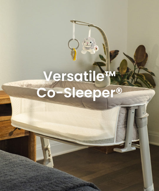 A Versatile Co-Sleeper bassinet in a nursery setting, featuring a white and gray fabric liner, a white metal frame, and an adjustable mobile with a plush bunny hanging from a curved arm.